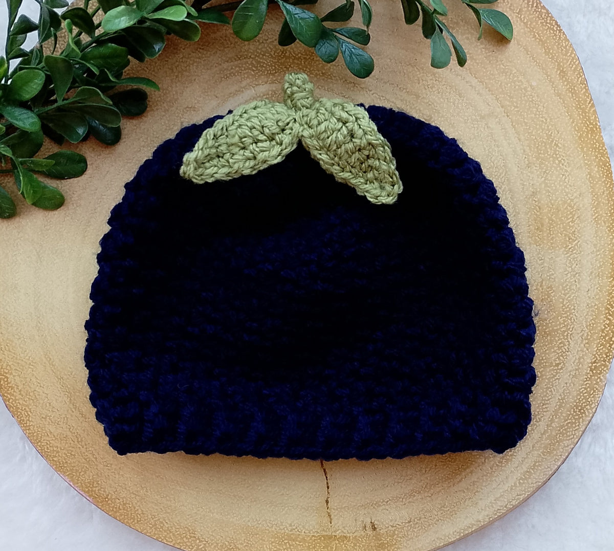A dark navy blue crocheted beanie in a blueberry hat style with a light green leaf applique on top, displayed on a round wooden slice with green leaves in the corner