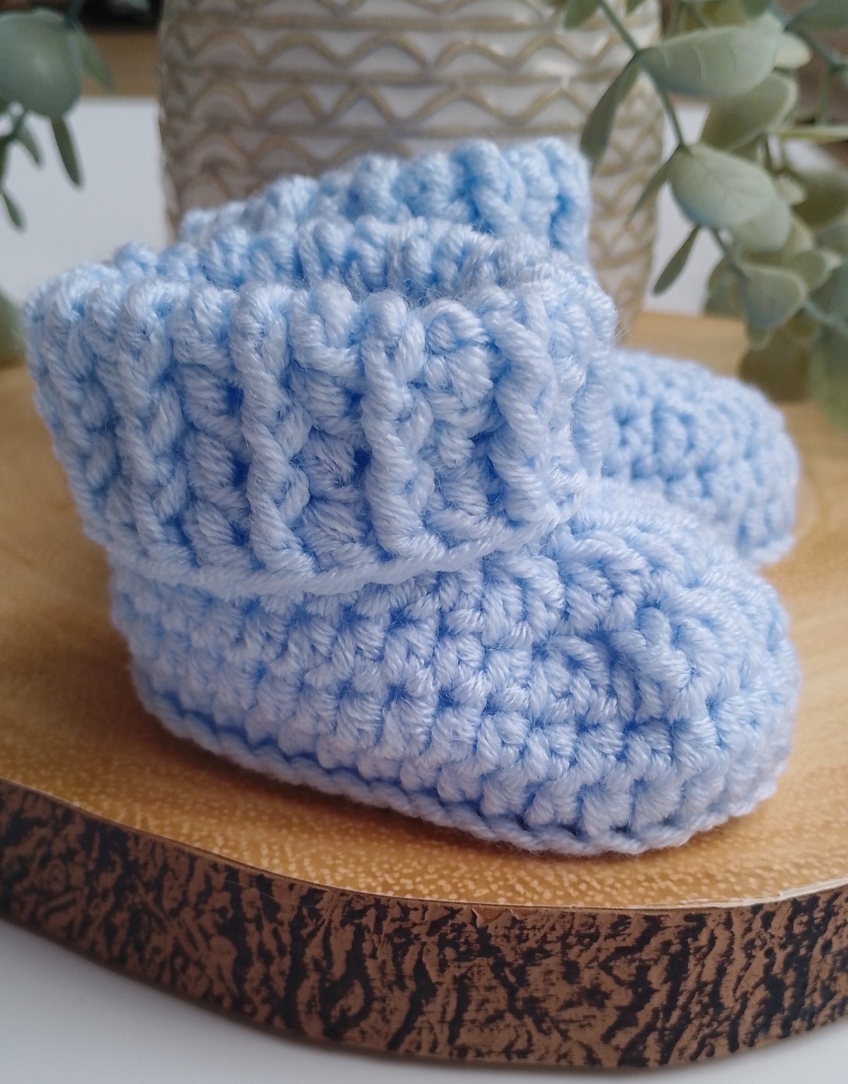 A pair of light blue crocheted baby booties displayed on a round wooden slice with a neutral patterned vase and green leaves in the background.