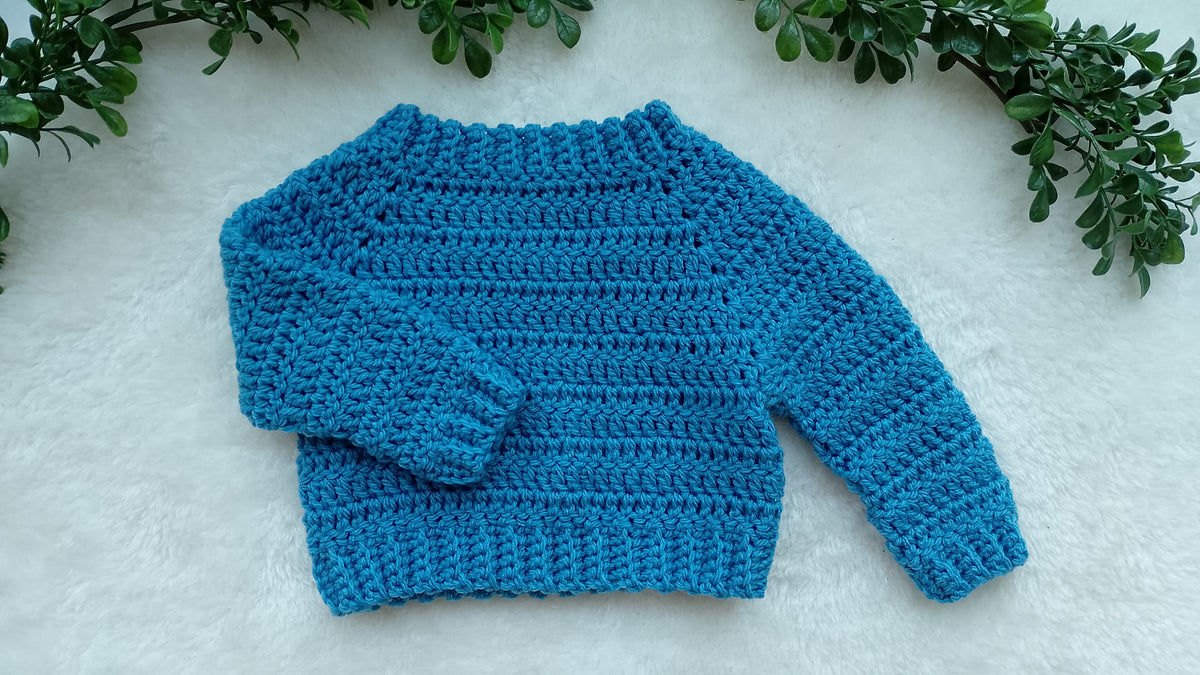 A cornish blue crocheted long-sleeve sweater laid flat on a white textured surface, framed by green leafy branches at the top corners.