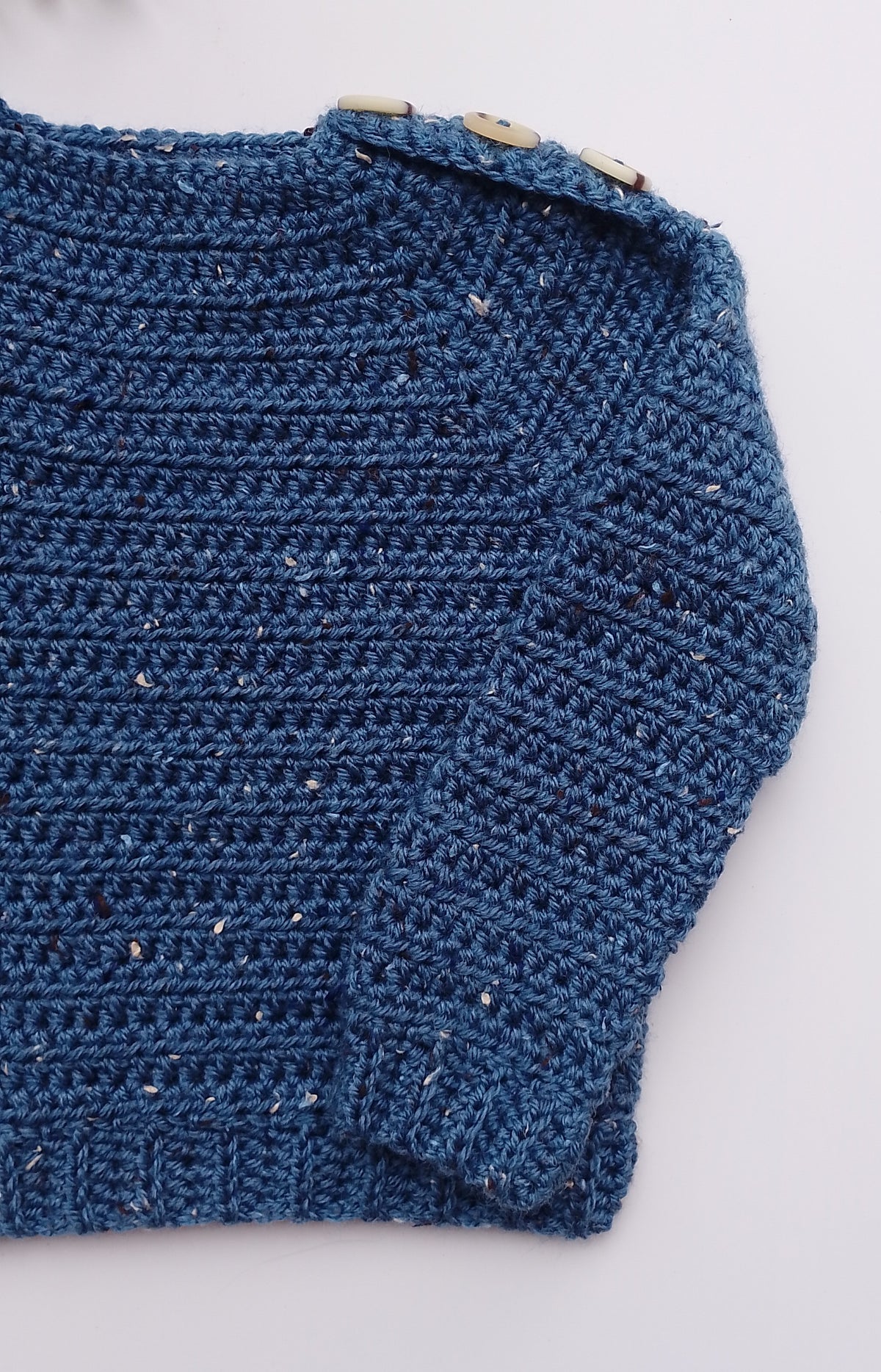 A close-up of a textured blue crocheted knit garment, showing the detailed stitch pattern and a small wooden button detail near the shoulder, laid flat on a light gray background.