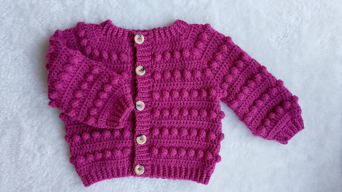 A raspberry pink crocheted cardigan with textured bobble stitches, long sleeves, and button closure, laid flat on a white textured background.