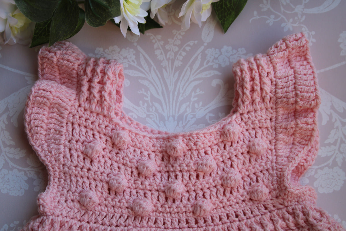 A close-up of a pink crocheted baby top or dress bodice with ruffled shoulder straps and textured bobble details, laid flat on a pale patterned surface with flowers at the top edge.