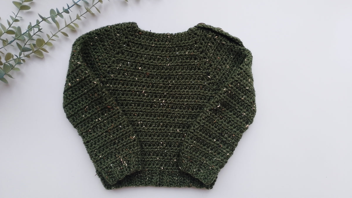 A dark green crocheted sweater with subtle gold flecks, laid flat on a white background with a decorative eucalyptus branch in the top-left corner.
