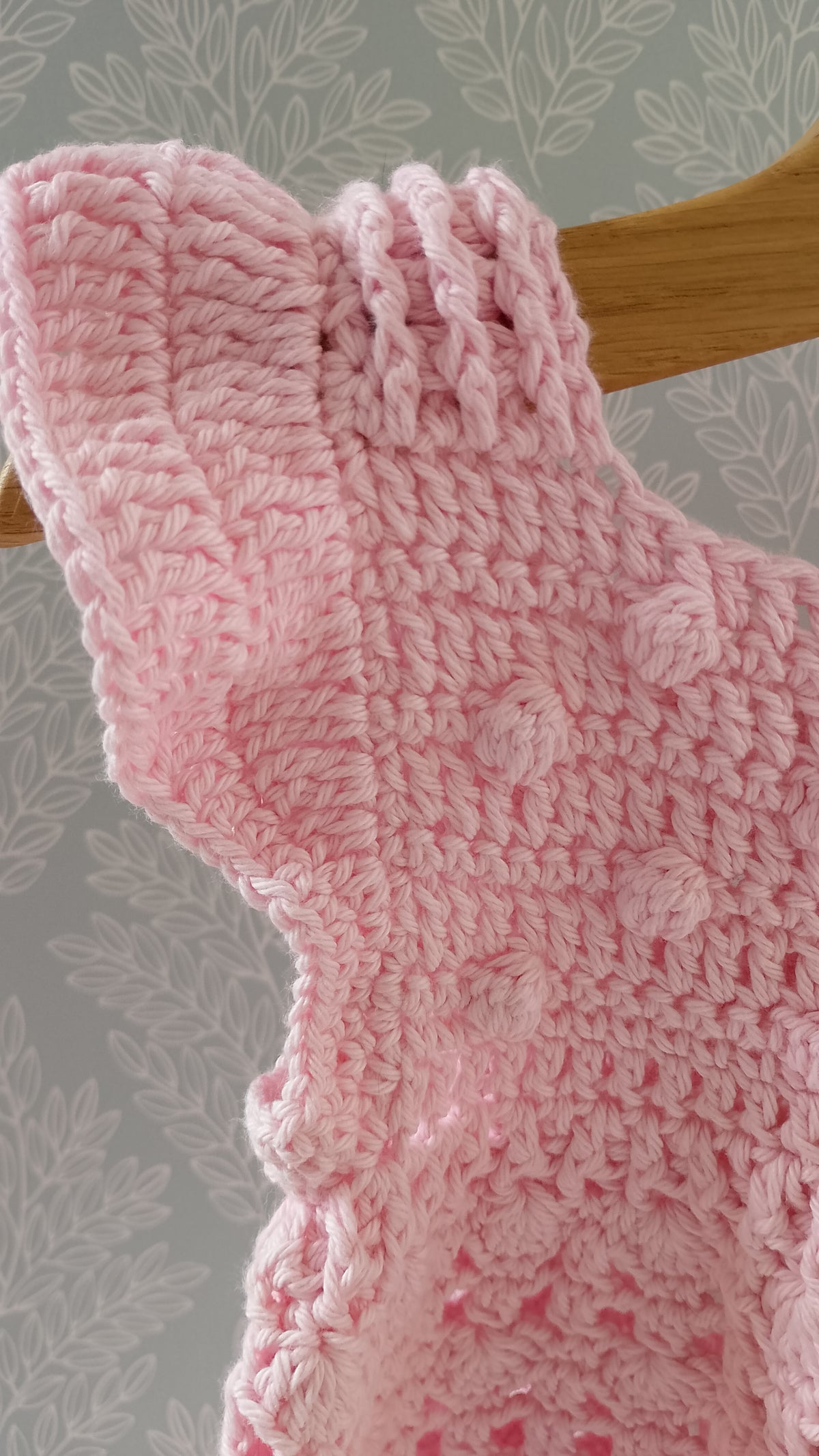 A close-up of a pink crocheted baby dress ruffled sleeve edge, hanging on a wooden hanger against a grey floral wallpaper background.