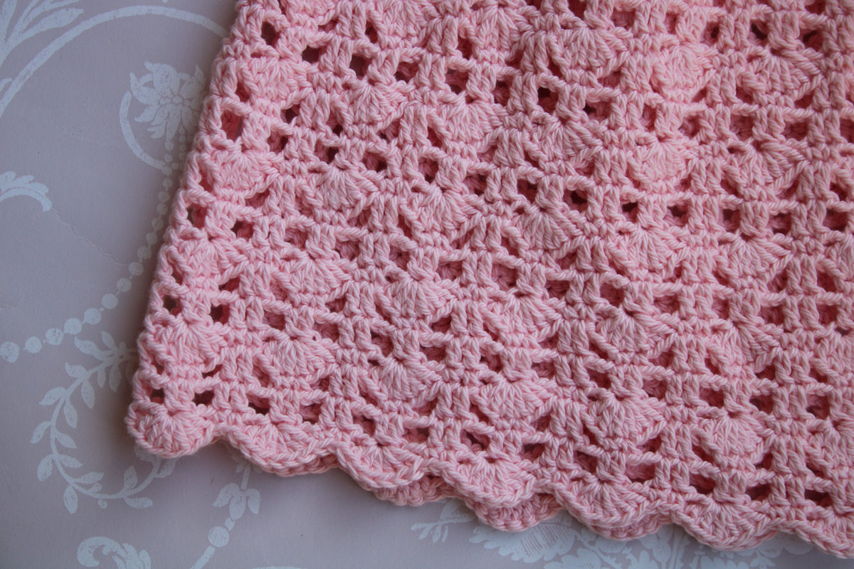 A close-up of a pink crocheted skirt with an openwork lace-like pattern and scalloped edge, laid on a pale patterned fabric background.