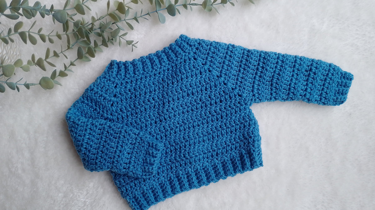 A bright blue crocheted baby sweater with long sleeves and a ribbed crew neck, laid flat on a white textured surface with green eucalyptus leaves in the corner.