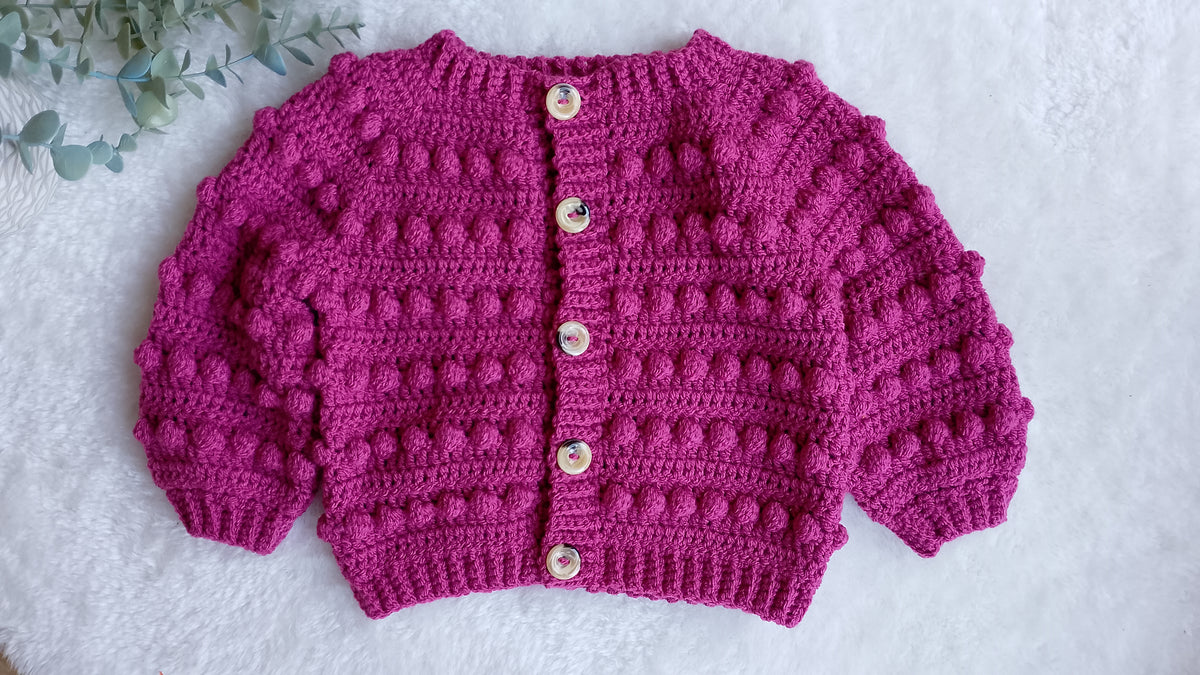 A pink crocheted cardigan with textured bobble details, featuring a high ribbed collar, button closure with beige buttons, and wide sleeves, laid flat on a white textured background.