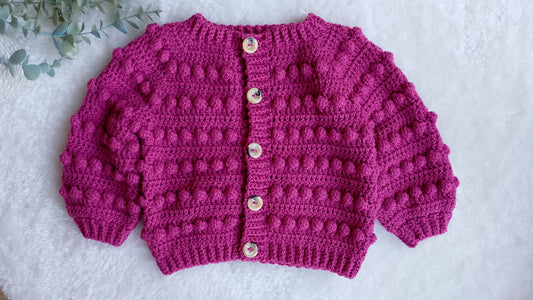 A pink crocheted cardigan with textured bobble details, featuring a high ribbed collar, button closure with beige buttons, and wide sleeves, laid flat on a white textured background.