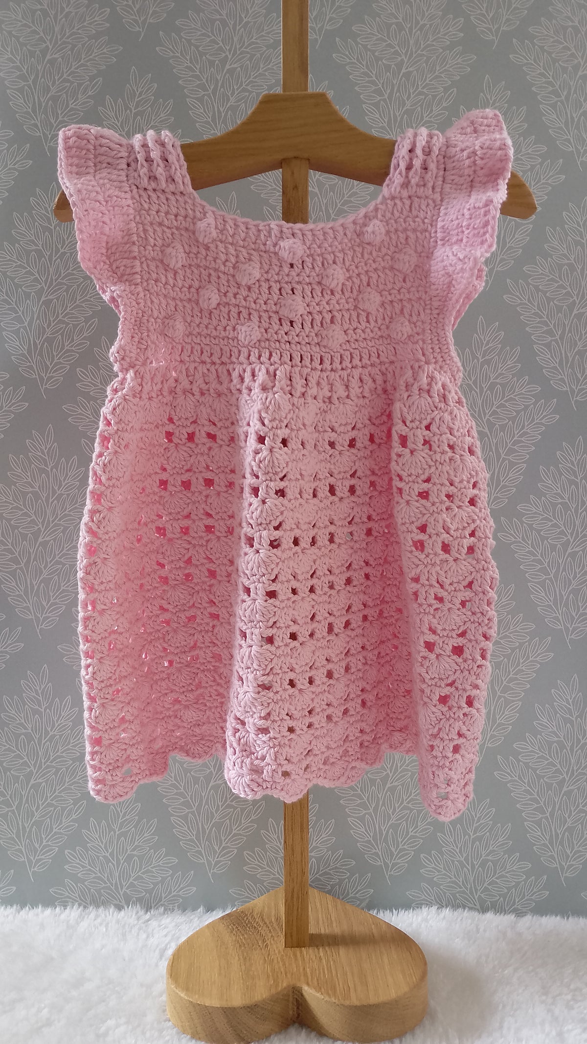 A pink crocheted baby dress with ruffled shoulder straps and a scalloped hem, displayed on a wooden hanger and stand against a gray patterned wall.