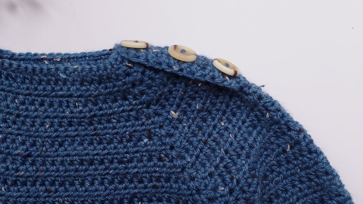 Close-up of a textured blue crocheted garment with three light beige buttons along the neckline, set against a plain white background.