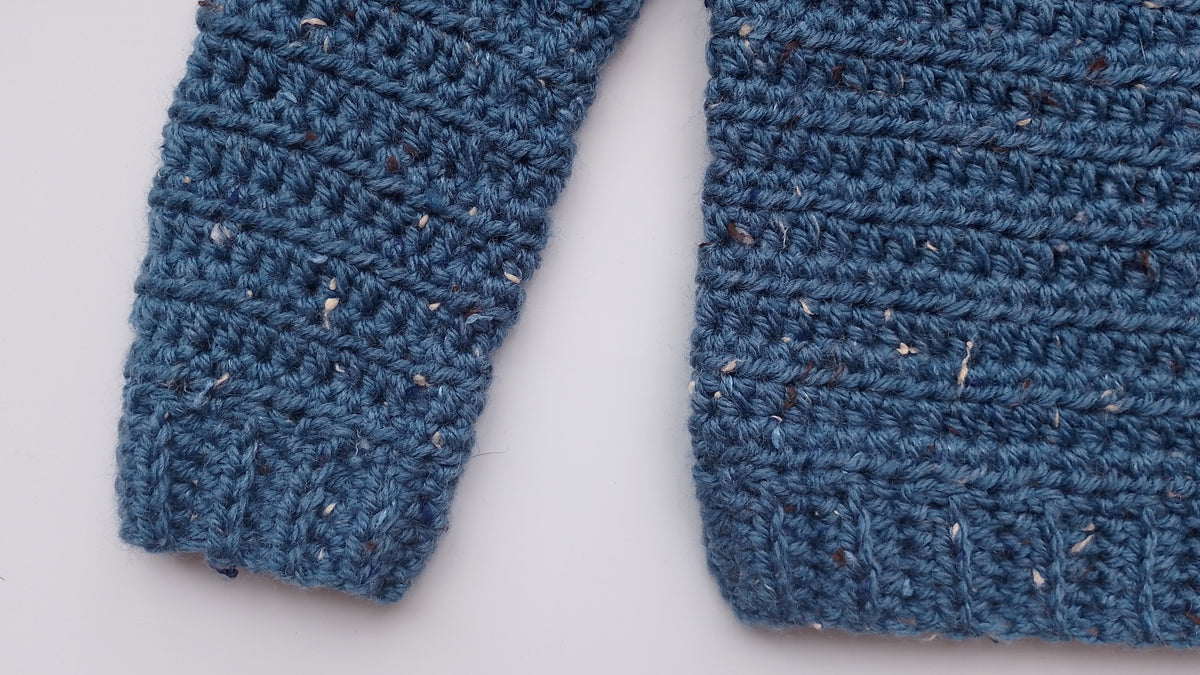 Close-up of a blue crocheted jumper, showing the textured knit pattern and flecks of lighter and darker yarn on a white background.