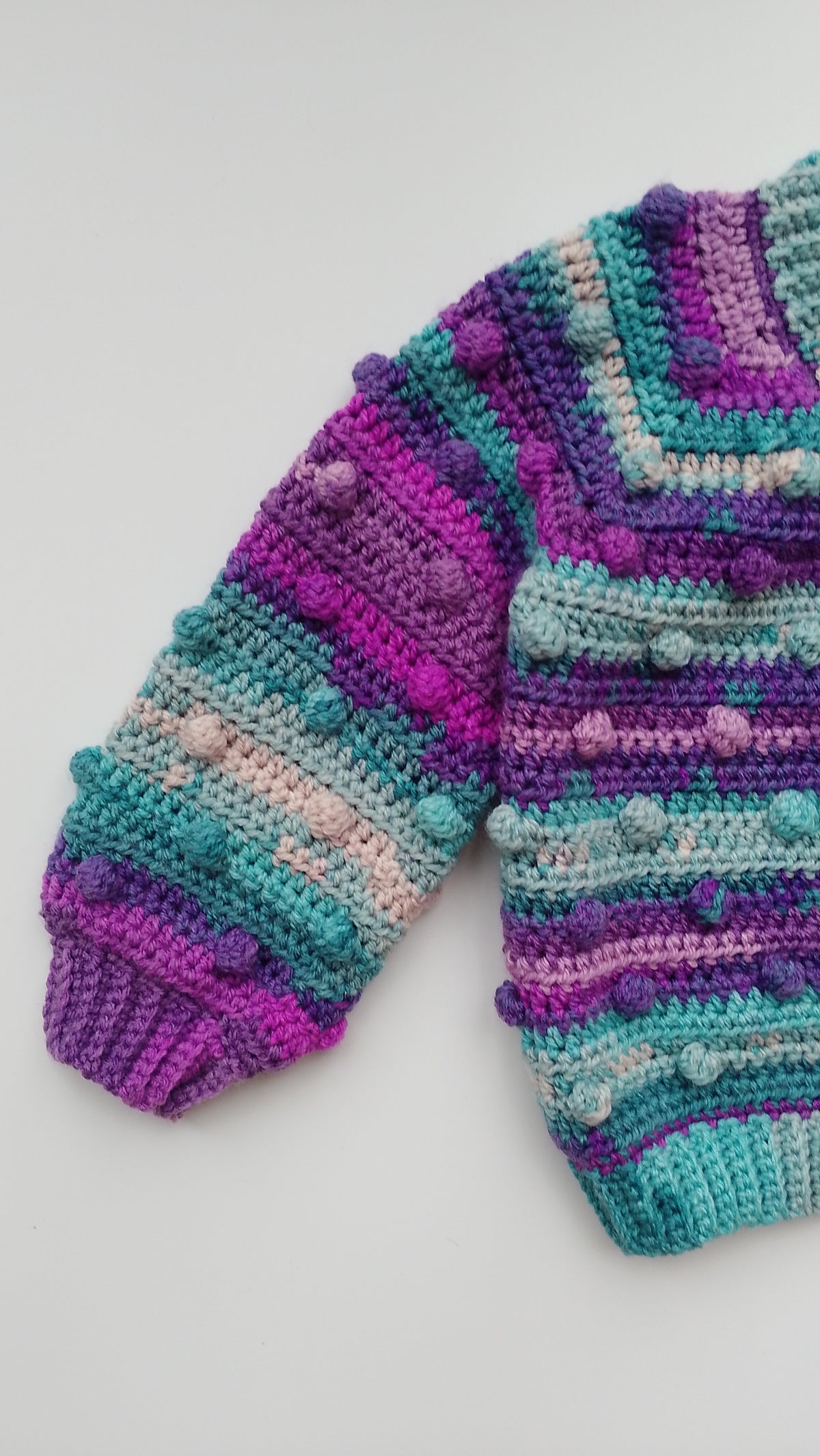 A close-up of a hand-crocheted knit sweater featuring horizontal stripes of purple, teal and beige with raised pom-pom details, laid flat on a light gray background.