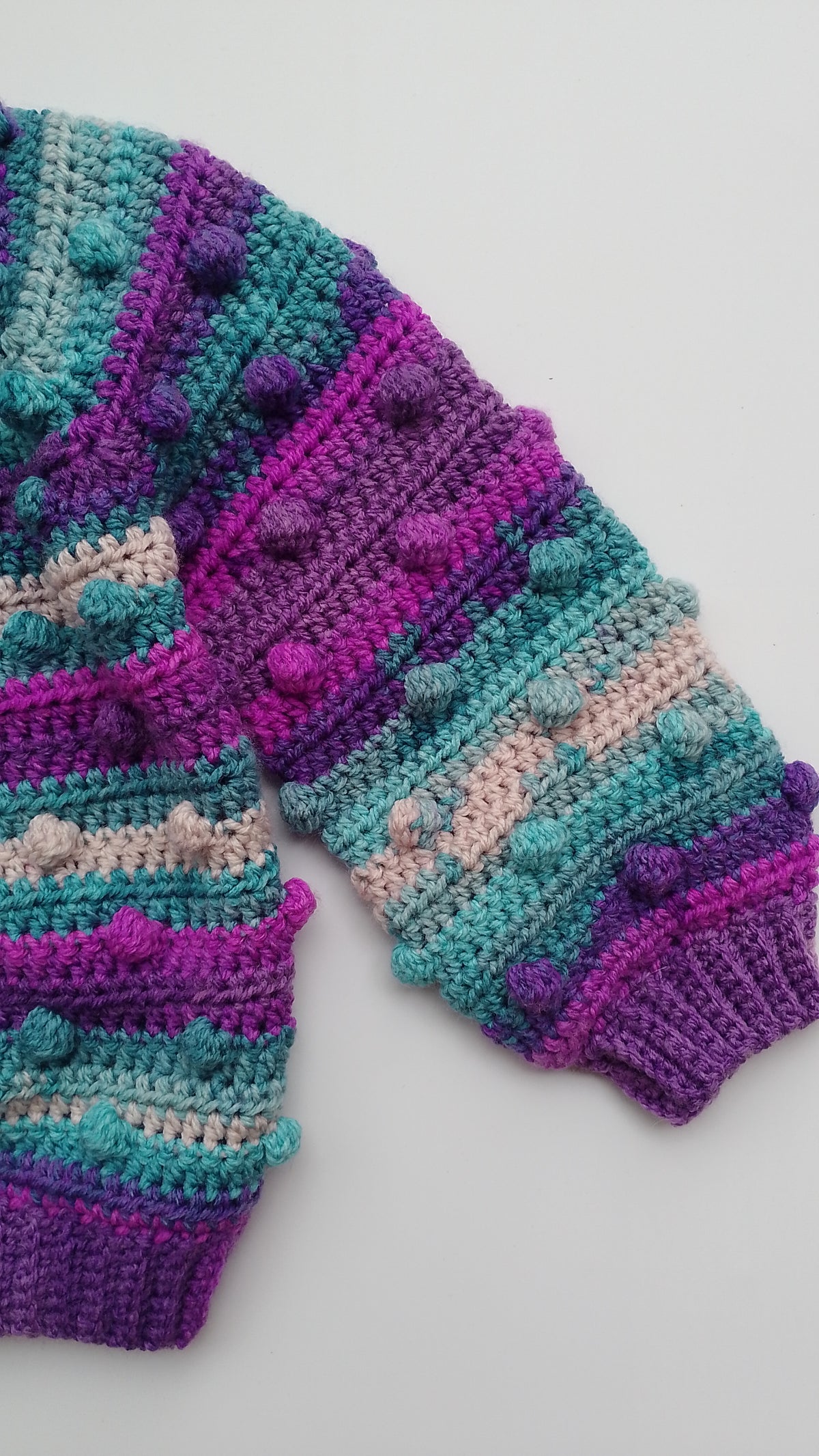 A close-up of a hand-crocheted knit sweater featuring textured rows of pom-poms, striped with teal, purple, magenta, and light pink yarn, laid flat on a light gray background.