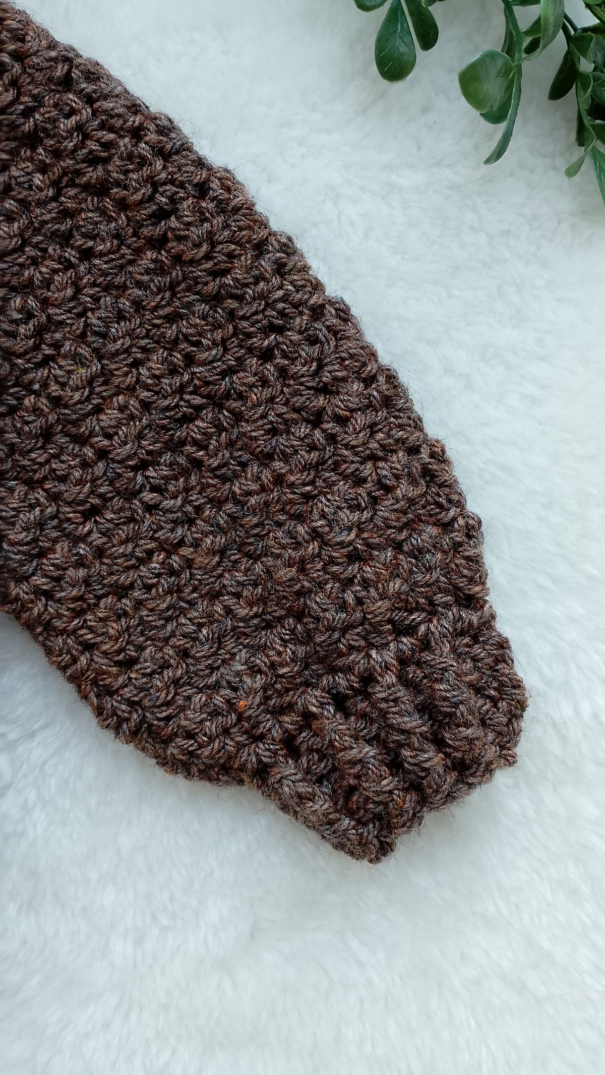 A close-up of a dark brown textured crochet sleeve and cuff laid on a white faux-fur surface with a small green plant leaf in the corner.