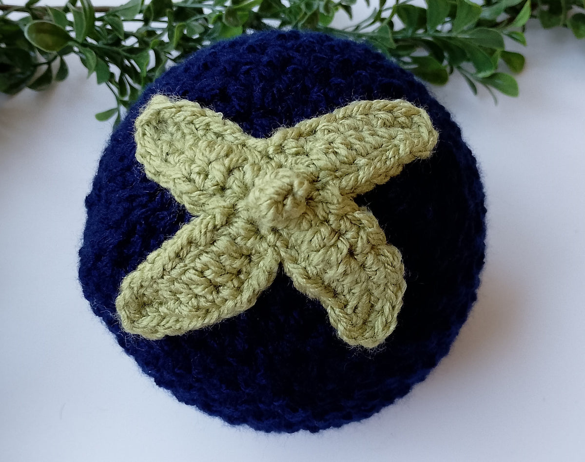 A navy blue crocheted blueberry hat with a textured stalk and leaves design in light olive green on top, displayed on a white surface with green foliage in the background.