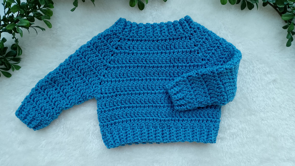 A bright blue crocheted long-sleeve sweater laid flat on a white textured surface, with green leaves framing the top corners.