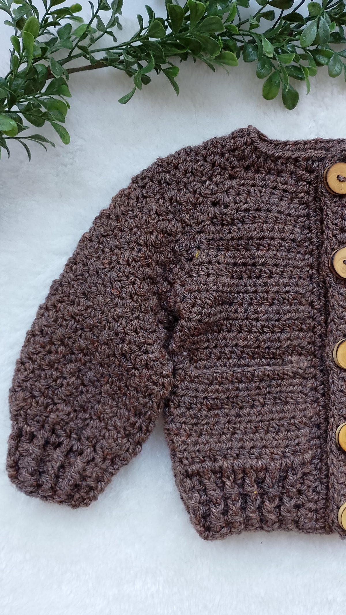 A brown crocheted cardigan sweater with wooden buttons, laid flat on a white textured surface with green leaves at the top.