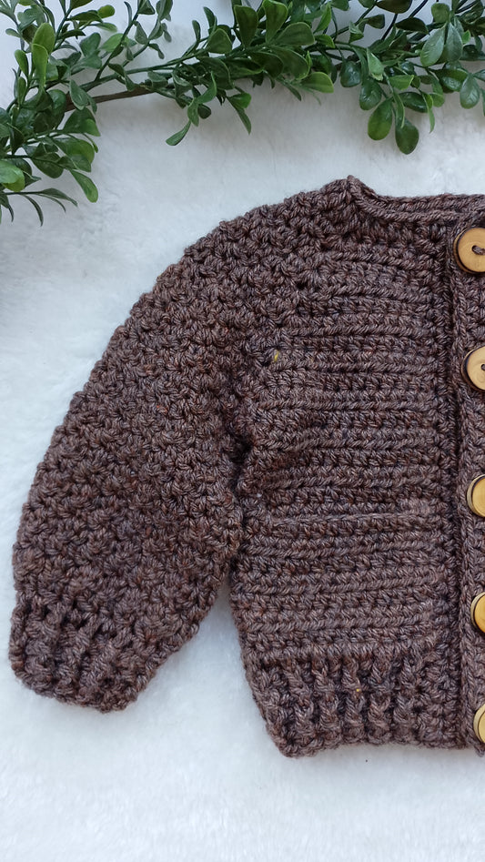 A brown crocheted cardigan sweater with wooden buttons, laid flat on a white textured surface with green leaves at the top.