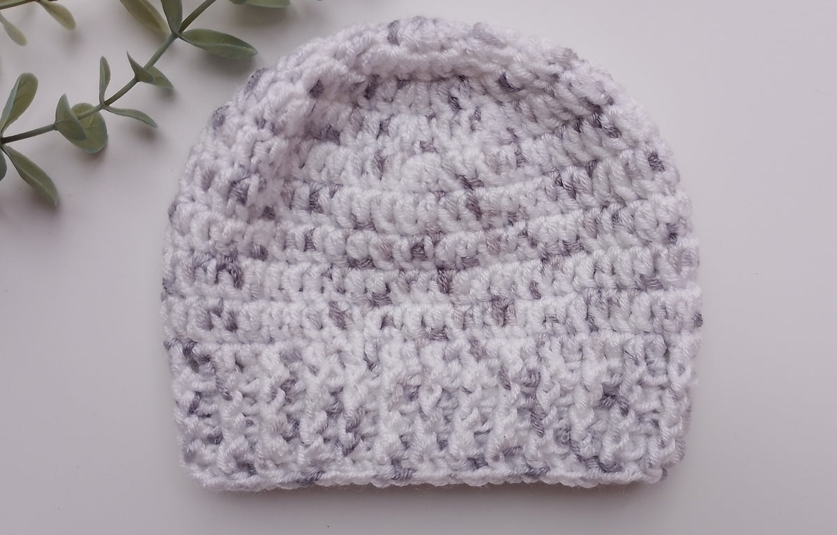 A light gray and white marled crochet beanie hat, photographed from a flat-lay perspective on a light gray background with a decorative green leafy sprig in the top-left corner.