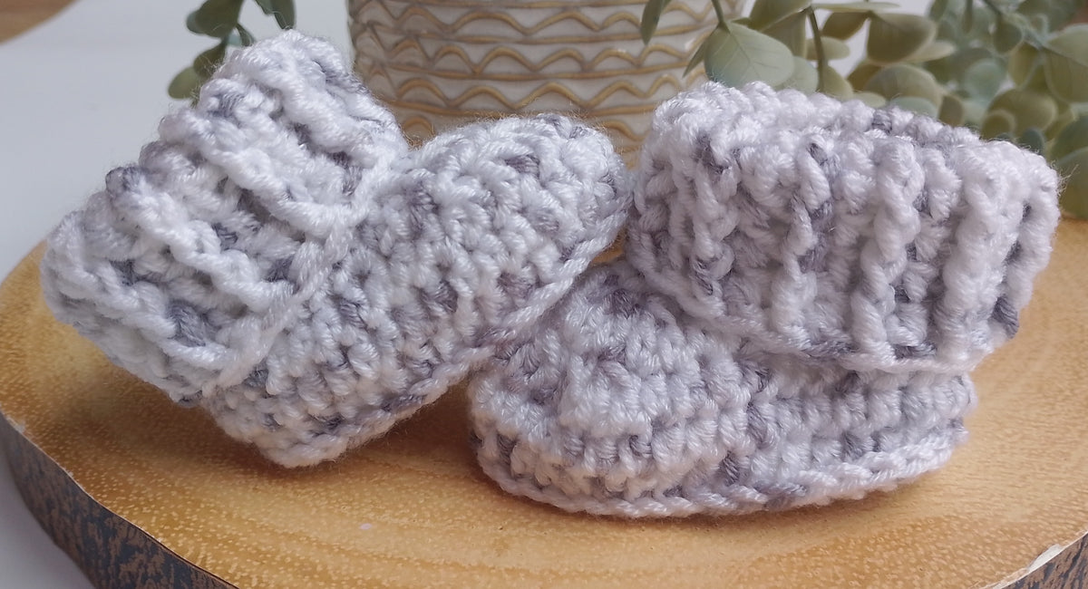 A pair of crocheted baby booties in a light grey and white variegated yarn, displayed on a round wooden slice with a neutral background.