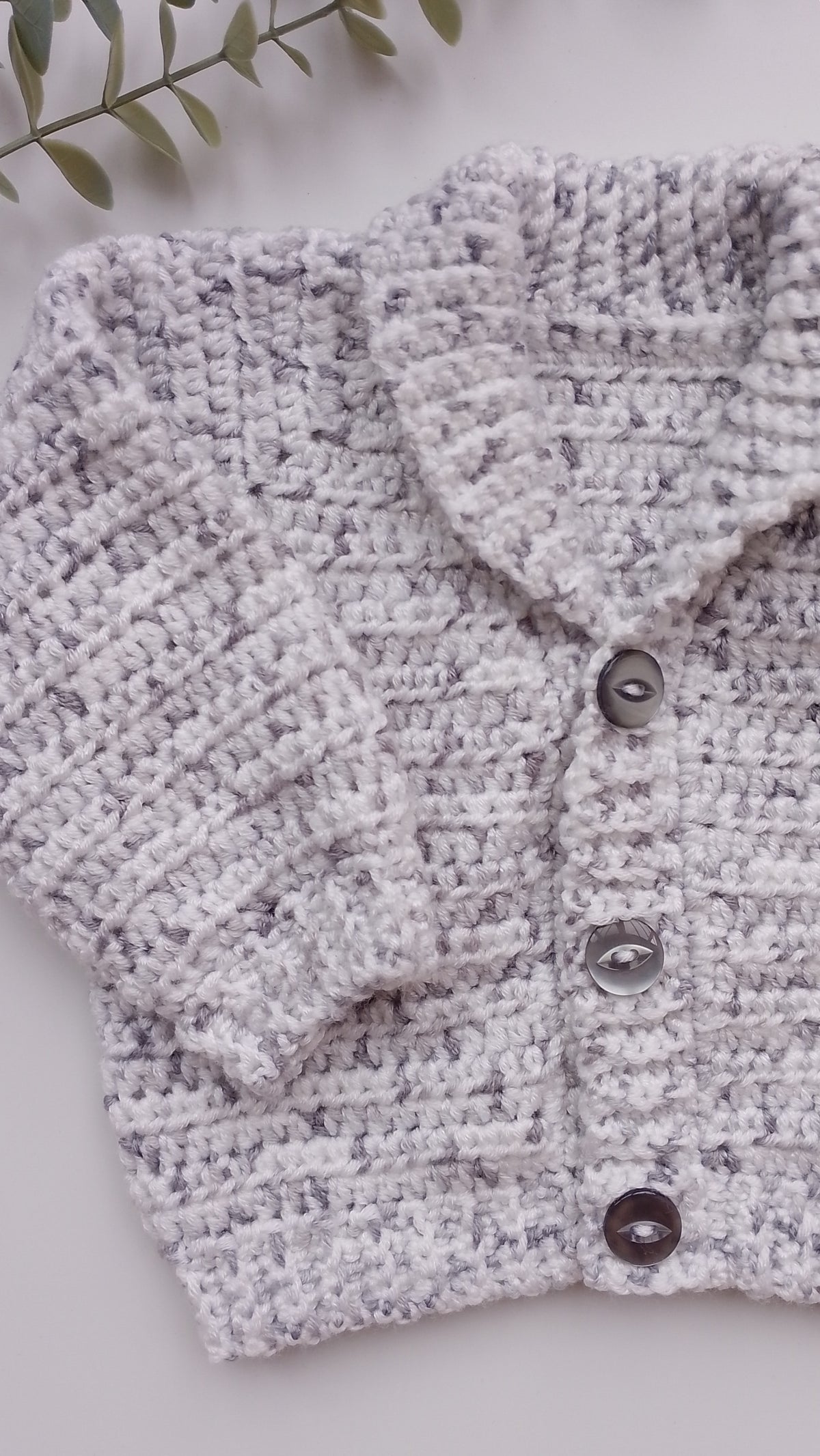 A close-up of a hand-crocheted cardigan in a heathered light gray and white yarn, featuring a collar and three dark brown buttons, laid flat on a light surface with a green leafy sprig in the corner.