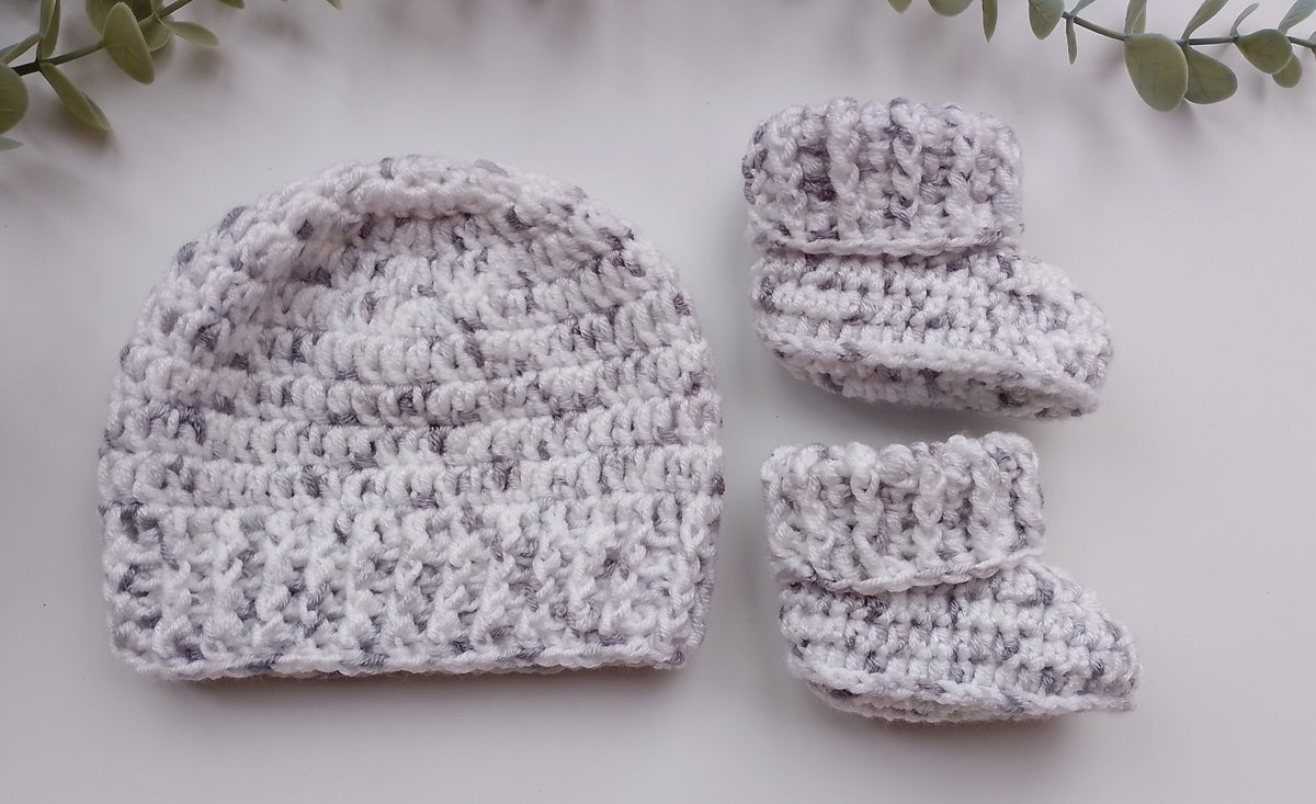 A handmade crochet baby hat and bootie set in a marled white and light gray yarn, styled flat-lay on a white surface with green leaves in the corners.